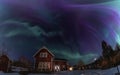 Panorama of Northern Lights in the clear starry skies shines above Swedish Wooden red house in countryside landscape. Green and