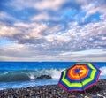 Rainbow umbrella on summer beach of tropical sea with azure waves and white ship under dramatic cloudy sky Royalty Free Stock Photo