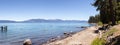 Panoramic View Of Rocky Beach And Dock At The Lake Surrounded By Mountains And Trees.