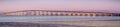 Panoramic view of San Mateo Bridge at sunset; electricity towers and power lines visible behind it; the San Mateo Bridge is