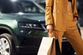 With paper bag in hand. Young fashionable woman in burgundy colored coat at daytime with her car