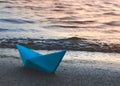 Paper blue ship is thrown on sandy shore of lake at sunset. Royalty Free Stock Photo