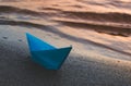 Paper blue ship is thrown on sandy shore of lake at sunset. Royalty Free Stock Photo