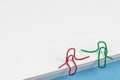 Paper clips red and green sitting on a white paper ream and talking or having a conversation. A photo with a copy space. Good for