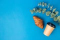 Paper coffee cup, croissant and eucalyptus leaves on Blue background. Good Morning or food concept. Flat lay banner, top view, Royalty Free Stock Photo