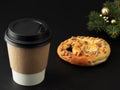 Paper cup with a hot drink, coffee or tea on a black background. Near a fresh pretzel and a Christmas tree. Close-up. Horizontal