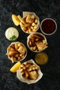 Paper cups with fried fish and chips, and sauces on black smokey background