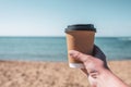 Paper mug, cup of coffee in woman hand, sand of sunny beach opposite the sea wave. summer. take away. breakfast.