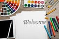 Paper with word Welcome, colorful markers, watercolor paints, palette and tablet on wooden table, flat lay