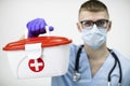 Paramedic in mask, glasses and blue latex gloves holds red cross medical case