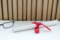 A parchment diploma scroll, rolled up with red ribbon beside a stack of books on white background.