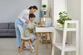 Mother helping son who is sitting at desk with laptop and doing homework in his notebook Royalty Free Stock Photo