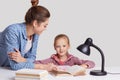 Parenthood, studying and education concept, Attractive blue eyed female child sits at workplace, reads book together with mother,