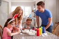 Parents With Children Wearing Bunny Ears Sitting At Table Decorating Eggs For Easter At Home