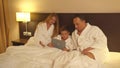 Parents and little son in sit on bed and watch something on tablet in hotel room