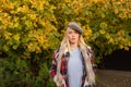 Parisian fashion style. Stylish blonde girl wearing plaid scarf and cap at fall autumnal yellow leaves background