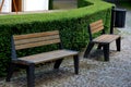 Park benches in front of a trimmed hedge. The benches are metal modern with wide black legs. The wood is brown natural. Granite