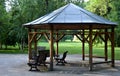 In the park there is a hexagonal gazebo with a metal roof in the shape of a pyramid. there are benches under the pergola. nature p