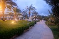 Park zone in empty resort place in the seaside with hotel buildings and palm trees near walk path in low season with night lightin