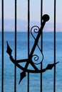 Part of wrought iron fence with silhouette of old antique vintage handmade anchor with decorations and light blue sea in back Royalty Free Stock Photo