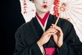 View of beautiful geisha in black kimono with red flowers in hair holding traditional asian umbrella isolated on black Royalty Free Stock Photo