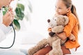 View of pediatrist in white coat and kid with teddy bear in clinic