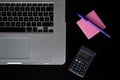 Partial view of a silver keyboard of a laptop with a calculator, and a pink post-it notes with a pencil on a dark wood desk