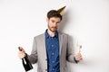 Party guy standing in birthday hat and celebrating, holding champagne bottle and glass, looking drunk, wearing grey suit