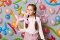 Party preparation. Birthday celebration. Cute little girl with braids blowing balloon standing against gray wall decorated with Royalty Free Stock Photo