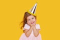 Party time. A joyful cute little child girl in a festive cap and elegant dress celebrates her birthday on a yellow
