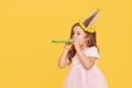 Party time. A joyful little girl in a festive cap and elegant dress celebrates her birthday. Blowing a whistle on a yellow