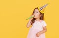 Party time. A joyful little girl in a festive cap and elegant dress celebrates her birthday. Blowing a whistle on a yellow