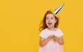 Party time. A joyful little girl in a festive cap and elegant dress celebrates her birthday. Blowing a whistle on a yellow Royalty Free Stock Photo