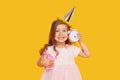 Party time. A joyful little girl in a festive cap and elegant dress celebrates her birthday. Holds a small alarm clock Royalty Free Stock Photo