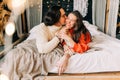 Passionate romantic couple spending home with garlands. Young woman and man are lying on bed in sweaters and hugging