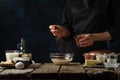 Pastry chef pours cocoa into glass bowl. Backstage of cooking waffle on rustic wooden table with ingredients on dark blue Royalty Free Stock Photo