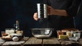 Pastry chef sifts flour into glass bowl on wooden table with ingredients for cooking on dark blue background. Backstage of Royalty Free Stock Photo