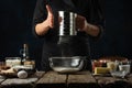 Pastry chef sifts flour into glass bowl on wooden table with variety of ingredients on dark blue background. Backstage of Royalty Free Stock Photo