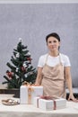 Pastry chef woman in an apron stands in kitchen next to gift boxes against Christmas tree background. Vertical frame