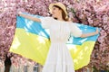 Patriotic Young Girl with Ukrainian Flag on Shoulders Smiling While Standing Outside near Sakura Blooming Trees. Proud