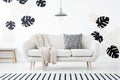 Patterned pillow and blanket on sofa in white living room interior with monstera leaves. Real photo