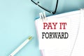 PAY IT FORWARD text on a sticky on notebook with pen and glasses , blue background Royalty Free Stock Photo
