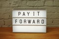 Pay It Forward word in light box on white brick wall and wooden background Royalty Free Stock Photo