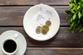 Pay restaurant bill by cash. Reciept and coins on plate on dark wooden background top view