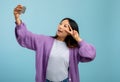 Peace selfie. Playful asian girl taking self-portrait on smartphone and showing v-sign, blue background with free space Royalty Free Stock Photo