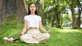 Peaceful Asian female sitting in lotus pose, meditating under the tree in the green park Royalty Free Stock Photo