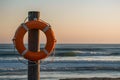 Peaceful beach scene at sunset with orange lifebuoy, wooden post, waves, and colorful sky Royalty Free Stock Photo