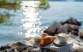 Peaceful breakfast setup with coffee and croissant on a lakeside rock, glistening water.