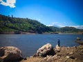 Peaceful Lake Water Dam Scenery With A Man Fishing Between Chunks Of Rock On A Sunny Day