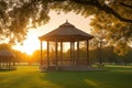 A peaceful park scene with a gazebo and trees in silhouette against the setting sun.